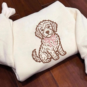 May include: Cream-colored sweatshirt featuring an embroidered brown and white dog wearing a pink bow. The dog is sitting with its tail curled. The sweatshirt has ribbed cuffs and a crew neck.