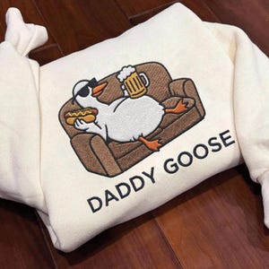 Könnte beinhalten: Cremefarbenes Sweatshirt mit einem gestickten Design einer Gans mit Sonnenbrille, die einen Hotdog und Bier hält und auf einem braunen Sofa liegt. Der Text "DADDY GOOSE" ist darunter aufgedruckt.