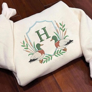 May include: A cream-coloured sweatshirt with an embroidered design. It features two ducks, cattails, and a green letter "H" inside a shield. The ducks have green heads, red chests, and grey wings. The design is framed by green leaves.