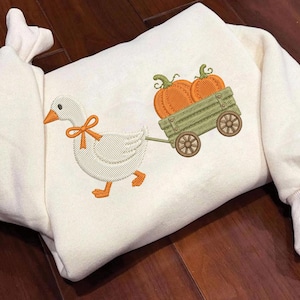 May include: A cream-colored long-sleeve sweatshirt featuring an embroidered design of a white goose with an orange bow around its neck, pulling a light green wooden wagon filled with two orange pumpkins with green stems and textured details.