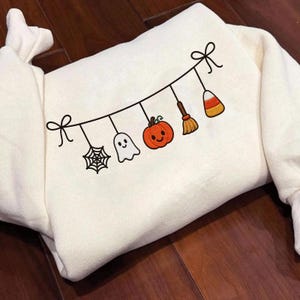 May include: A cream-colored sweatshirt featuring a black line-art illustration of a hanging garland. The garland displays a spider web, a white ghost, an orange pumpkin, a brown broom, and a piece of candy corn, all tied with bows at each end.