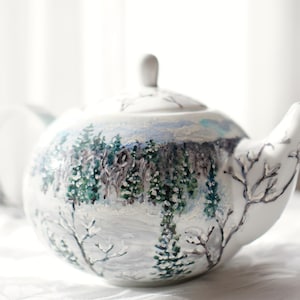 Puede incluir: Tetera de cerámica blanca con un diseño de paisaje invernal. La escena pintada a mano presenta árboles cubiertos de nieve y una cordillera en tonos azules, verdes y blancos. La tetera tiene un pico curvo y un asa pequeña en la tapa.