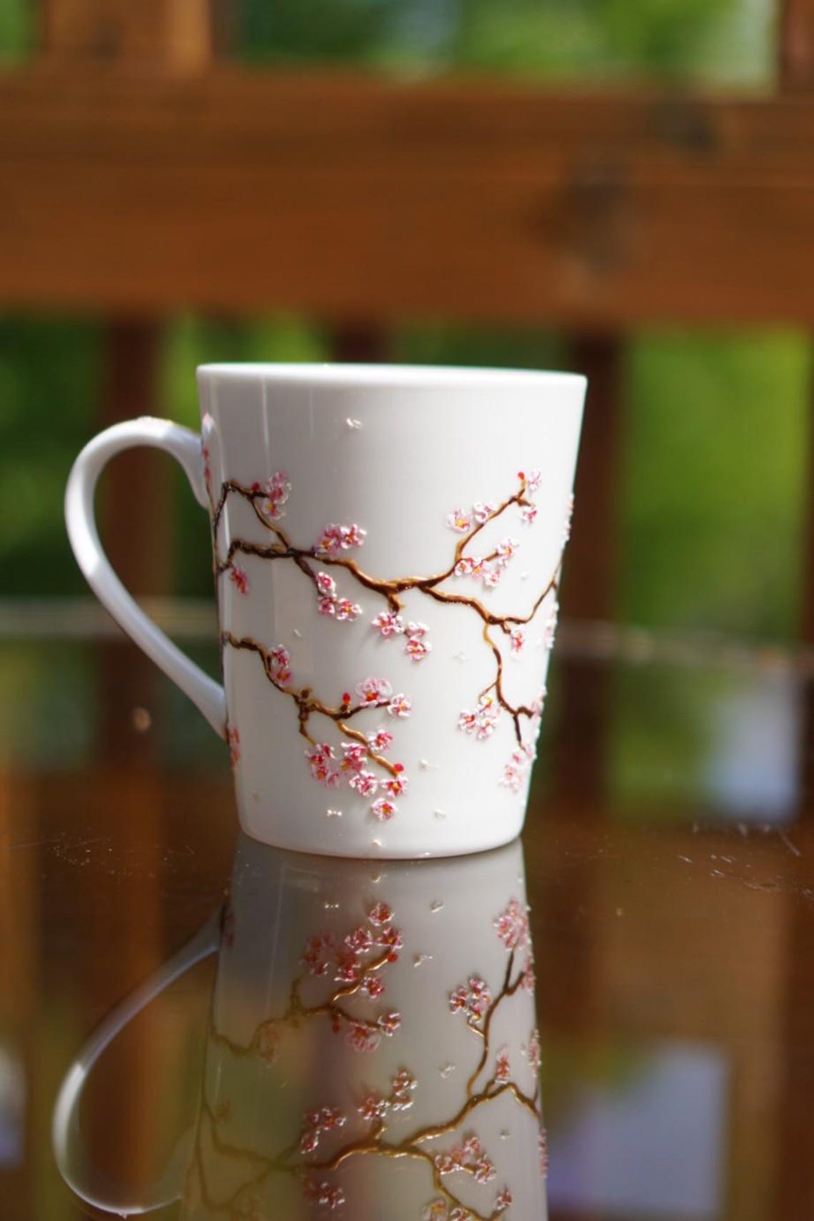 May include: White ceramic mug with a curved handle, adorned with a delicate design of pink cherry blossom branches. The branches are brown and golden, and the flowers are a delicate pink and white. The branches and flowers are sculpted on the surface of the mug.