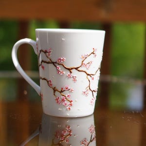 May include: White ceramic mug with a curved handle, adorned with a delicate design of pink cherry blossom branches. The branches are brown, and the flowers are a vibrant pink. The mug is reflected on a glossy surface.