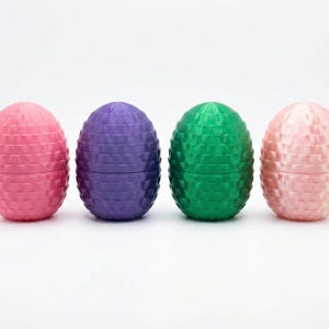 Op de afbeelding: Vier eivormige containers op een rij. De kleuren zijn roze, paars, groen en lichtroze. Elke container heeft een gestructureerd, geometrisch patroon. De containers zijn gesloten en staan op een witte ondergrond.