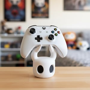 May include: A white Xbox controller rests on a white, stylized character-shaped stand. The controller has black buttons and colored action buttons. The stand has large, black eye-like openings. The background is blurred, with posters and shelves visible.