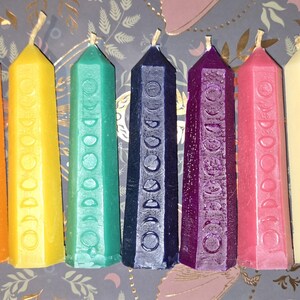 Peut inclure: Sept bougies colorées en forme d'obélisque, allant du rouge au noir. Chaque bougie est ornée d'un motif de phases lunaires sculptées. Ces bougies décoratives sont disposées en rangée, présentant un spectre de couleurs.