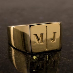 Può includere: Un anello a sigillo dorato con le iniziali incise "M" e "J". L'anello ha una forma rettangolare con bordi arrotondati e una finitura lucida. Le iniziali sono separate da una linea verticale.