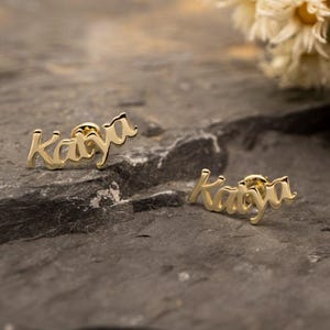 Puede incluir: Dos pendientes dorados que deletrean el nombre "Katya" en una fuente cursiva. Los pendientes se muestran sobre una superficie oscura y texturizada. Los pendientes están enfocados, con un fondo borroso.