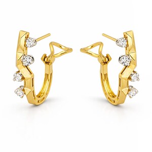 May include: A pair of gold-colored hoop earrings with a geometric design. Each earring features three clear, round gemstones. The earrings have a post and clasp closure. The earrings are displayed against a white background.