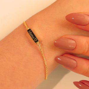 May include: A gold-colored bracelet with a black rectangular charm that reads "Mama" with a heart symbol. The bracelet is worn on a wrist and has a delicate chain design.