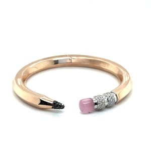 Puede incluir: Brazalete de oro rosa con forma de lápiz, con punta negra y goma rosa. El extremo de la goma está adornado con pequeñas piedras brillantes. El brazalete es abierto, con la punta del lápiz y los extremos de la goma enfrentados.