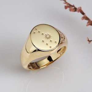 Peut inclure: Une bague chevalière en or avec un motif de constellation. La bague présente une face ovale polie avec une étoile filante et plusieurs petites étoiles. La bague est en métal doré brillant et possède un anneau lisse et arrondi.
