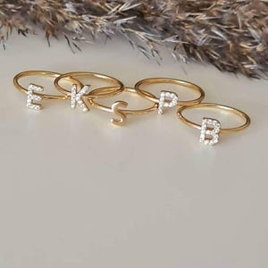 Puede incluir: Cinco anillos dorados, cada uno con una letra diferente: E, K, S, P y B. Las letras están incrustadas con pequeñas piedras preciosas transparentes. Los anillos se muestran sobre una superficie clara, con un fondo borroso.