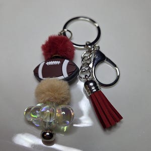 May include: A football-themed keychain with a brown and white football charm, red and tan fluffy pom-poms, iridescent beads, and a red tassel. The keychain has a silver keyring and clasp.