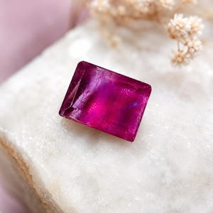 May include: A rectangular, faceted gemstone in a deep, vibrant magenta-pink hue. The stone features a polished, reflective surface with visible internal inclusions and sharp, clean edges, showcasing a translucent quality and a rich, saturated color.
