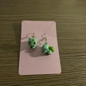 Könnte beinhalten: Ein Paar hellgrüne Bulbasaur-Ohrringe. Ein Ohrring zeigt den Kopf von Bulbasaur, der andere zeigt den Körper von Bulbasaur mit einer grünen Knolle auf dem Rücken. Die Ohrringe befinden sich auf einer lavendelfarbenen Karte.