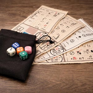 Puede incluir: Una bolsa negra con cordón que contiene seis dados multicolores, junto a varias hojas de juego beige con símbolos y texto impresos. Las piezas del juego están sobre una superficie de madera.