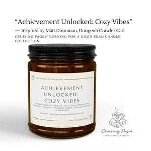 Op de afbeelding: Een bruine glazen kaars met een zwarte deksel en een wit etiket. Het etiket leest: Achievement Unlocked: Cozy Vibes, 100% Natural Soy Wax Blend, Congratulations, Crawler. The system approves this fragrance. Reward? A pleasant aroma. You have received +1 ambiance. Warning: Excessive relaxation detected. Cruising Pages.