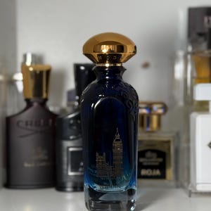 Pode incluir: Um frasco de perfume azul escuro com tampa dourada. A garrafa apresenta uma imagem gravada do Big Ben. A garrafa está em uma prateleira com outros frascos de perfume ao fundo.