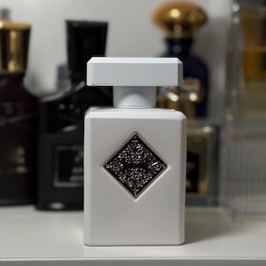 Könnte beinhalten: Eine weiße, rechteckige Parfümflasche mit einem silbernen und schwarzen Emblem. Die Flasche hat einen weißen Deckel und ist von anderen Parfümflaschen umgeben. Die Parfümflasche steht auf einer weißen Oberfläche.