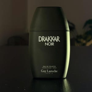 Pode incluir: Um frasco de spray de Eau de Toilette Drakkar Noir preto fosco. O frasco tem uma forma retangular arredondada com uma tampa preta. O nome da marca e do produto está em letras brancas. O logotipo da Guy Laroche está na parte inferior.