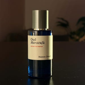 Pode incluir: Um frasco de perfume azul escuro com tampa prateada. O rótulo diz "Oud Maracujá Extrait de Parfum" e "Maison Cruel". O frasco está sobre uma superfície escura.