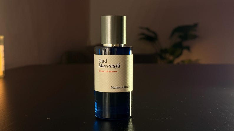 Puede incluir: Un frasco de perfume azul oscuro con tap&oacute;n plateado. La etiqueta dice "Oud Maracuj&aacute; Extrait de Parfum" y "Maison Cruel". El frasco est&aacute; sobre una superficie oscura.