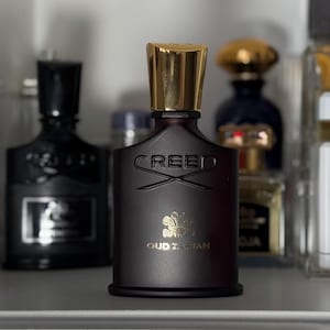 Puede incluir: Un frasco de perfume burdeos oscuro con una tapa dorada. La botella tiene la palabra "CREED" en relieve en la parte delantera, junto con las palabras "OUD ZARIAN" y un emblema dorado. Se ven otros frascos de perfume en el fondo.