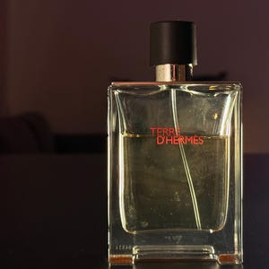 May include: A clear glass perfume bottle with a black cap and silver neck. The bottle has a rectangular shape and contains a light amber liquid. The words "TERRE D'HERMES" are printed in red on the front.