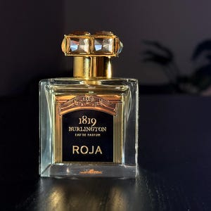 Pode incluir: Um frasco de perfume quadrado de vidro com tampa e detalhes dourados. O frasco tem um rótulo preto com letras douradas que diz "1819 Burlington Roja". Uma vela acesa é visível ao fundo.