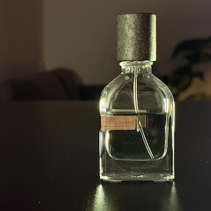 Puede incluir: Un frasco de perfume de vidrio transparente con una tapa oscura y texturizada. El frasco contiene un líquido de color claro y tiene una etiqueta que dice "ORTO PARISI". El frasco está sobre una superficie oscura.