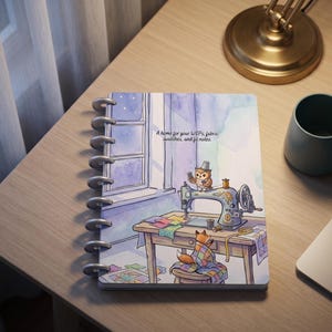 May include: A disc-bound notebook featuring a watercolor illustration of an owl sewing at a desk with a fox nearby. The cover includes the text: A home for your WIPs, fabric swatches, and fit notes. The scene depicts a window, a sewing machine, and colorful fabric scraps in soft purple and blue tones.