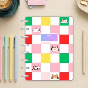 May include: A disc-bound notebook featuring a checkered pattern of pink, white, yellow, red, and green squares. Small cartoon cat illustrations are placed within some squares. A purple rectangular label with the word Notes is centered.