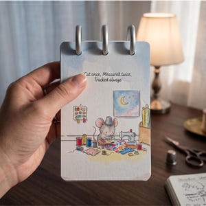 May include: A rectangular notepad with three silver rings at the top, featuring a watercolor illustration of a mouse sewing with colorful fabric scraps and spools of thread. The text "Cut once, Measured twice, Tracked always" is printed in black.
