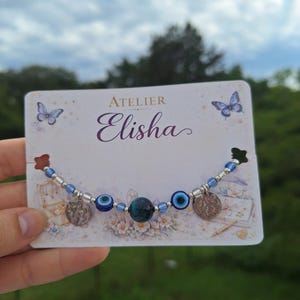 Peut inclure: Un bracelet de perles avec une pierre œil-de-tigre bleu foncé au centre, deux perles œil bleu plus petites, des intercalaires argentés et deux breloques circulaires texturées. Le bracelet est présenté sur une carte blanche Atelier Elisha.