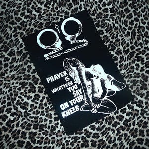 May include: A black rectangular patch featuring a white graphic print of handcuffs and a kneeling figure. The text reads PRAYER IS WHATEVER YOU SAY ON YOUR KNEES. The design is rendered in a high-contrast black and white style with detailed line work.