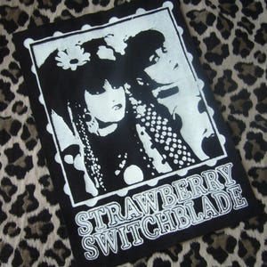 May include: Black and white patch featuring a graphic of two women with long hair and floral accents. The text "STRAWBERRY SWITCHBLADE" is printed in white block letters at the bottom of the patch. The patch is rectangular with a white border.