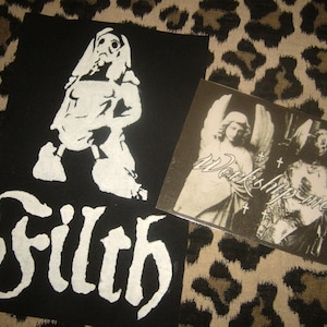 Op de afbeelding: Twee patches en een kleine vierkante kaart op een luipaardprint achtergrond. Eén patch is zwart met een witte illustratie van een figuur en het woord "Filth". De andere is een vierkante kaart met twee engel illustraties en de woorden "Worship me".