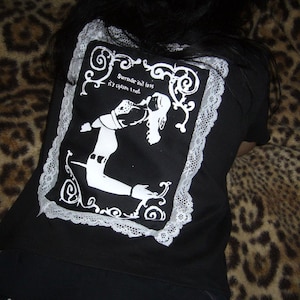 Op de afbeelding: Een zwart t-shirt met een rechthoekige grafische print op de achterkant. De print toont een wit silhouet van een figuur in een pose, omringd door sierlijke krullen en de tekst 'Surrender isn't loss, it's chosen trust', omlijst door witte kanten randen.