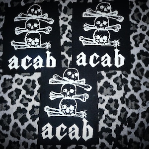 May include: Three rectangular black fabric patches featuring a white screen-printed graphic of a vertical stack of three skulls with crossed bones between them. Below the skulls, the text "acab" is printed in a bold, white, gothic-style font.
