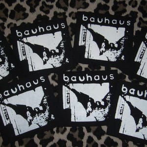 May include: A collection of black and white Bauhaus patches. Each patch displays the band's name in white, above a graphic design. The patches are square and set against a leopard print background.