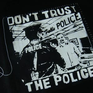 May include: A black t-shirt featuring a white screen-printed graphic of a police officer with a knife behind their back. The text reads DON'T TRUST THE POLICE at the top and bottom, with the word POLICE also printed on the officer's back.