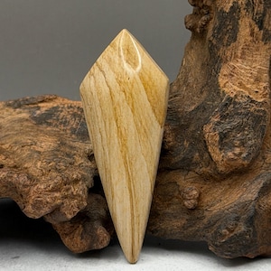 May include: A polished, elongated gemstone with a diamond-like shape. The stone features a warm, light brown color with natural wood grain patterns. The gemstone is displayed against a textured, dark brown wooden background.