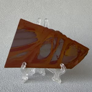 May include: A triangular, polished stone with a vibrant mix of orange, yellow, and gray hues. The stone is displayed on a clear plastic stand, showcasing its natural patterns and textures. The edges are rough, adding to its unique character.