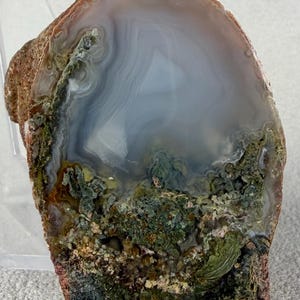 May include: A polished agate slice featuring a translucent, pale blue-grey center with delicate banding. The lower portion displays a textured, mossy green inclusion with earthy brown and dark green mineral deposits along the outer, rough edge.