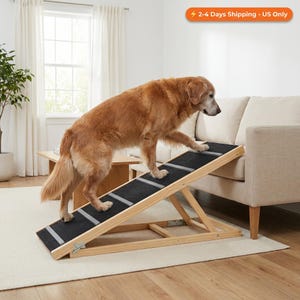Op de afbeelding: Een golden retriever loopt een houten dierenhelling op met een zwart, antislip oppervlak. De helling heeft zilveren strepen en staat naast een beige bank in een woonkamer. De tekst "2-4 Days Shipping - US Only" is zichtbaar.