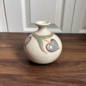 May include: A small, off-white ceramic vase with a round body and a narrow neck. The vase is decorated with blue and teal heart-shaped designs and gold accents. The vase is approximately 5 inches tall.