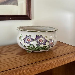 May include: A white ceramic planter with a scalloped rim and three small feet. The planter is decorated with purple and green floral designs and a green border. The planter is sitting on a wooden surface.