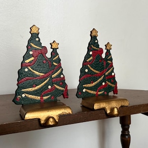 May include: A pair of metal stocking holders shaped like Christmas trees. Each tree is painted dark green with gold garland, red ribbons, and white dots. The trees are topped with gold stars and sit on solid gold-colored rectangular bases.